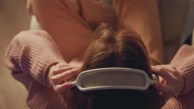 Top-down close-up of person seated on upholstered chair, placing white over-ear headphones and adjusting earcups with both hands while head tilts slightly back, creating calm listening moment in