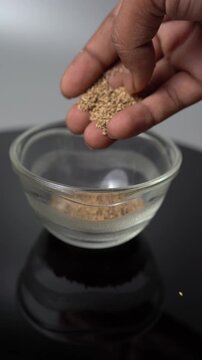Close-up of a person's hand pouring carom seeds (Ajwain) into a clear glass bowl, showcasing cooking preparation