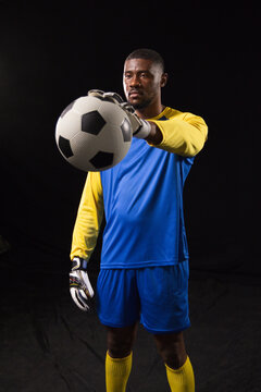 African American man standing in studio wearing blue goalkeeper kit and gloves holding soccer ball