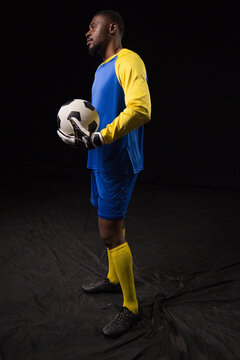Mid adult goalkeeper standing in studio in blue jersey holding black white soccer ball with gloves