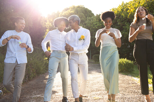 Diverse friends walking on gravel path, blowing bubbles, showing boutonniere, wearing smart clothes