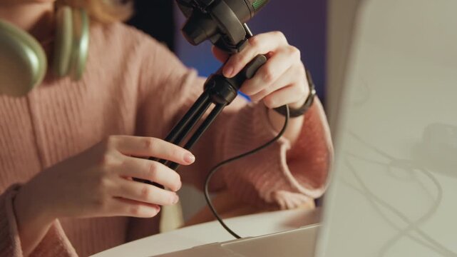 Close-up of person tightening and positioning RGB-lit condenser microphone on small desktop tripod next to computer monitor, fingers rotating mount knob while braided cable rests nearby