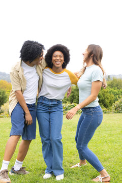 Diverse friends standing with arms around shoulders on grassy slope, smiling and wearing jeans