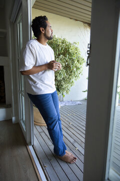 Man standing in doorway to covered deck, holding ceramic mug, barefoot and wearing white T-shirt