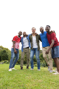 Diverse male friends linking arms, laughing on grass in park, wearing casual shirts and sneakers