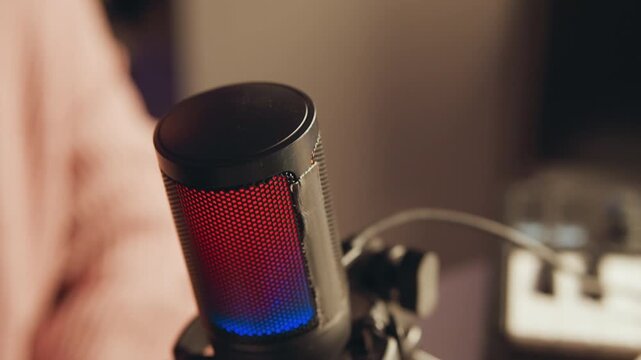 Tight angled view of large condenser microphone with metal mesh grille and foam top, glowing red and blue beside partially visible presenter gesturing during recording in home studio