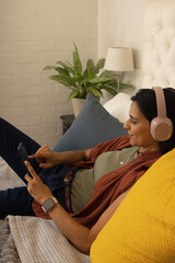 Indian woman reclining on bed in bedroom, using tablet with headphones and smartwatch, copy space © wavebreak3