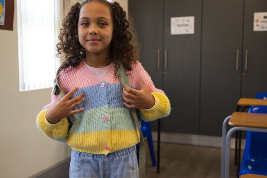 African American girl child standing at classroom front holding backpack straps, in pastel cardigan