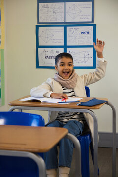 African American girl raising hand and smiling while sitting at desk in classroom with red pencil