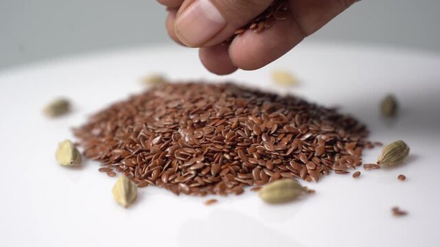 Close-up of a hand sprinkling golden flax seeds onto a white surface with green cardamom pods scattered around