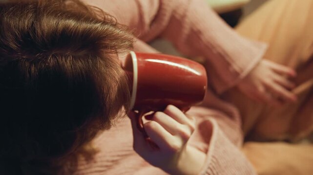 Oblique overhead close-up shows seated person lifting red ceramic mug, wrist raised and fingers wrapped around handle, creating feeling of comfort, calm morning routine, and quiet indoor warmth