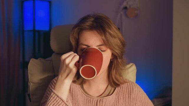 Young woman in neutral upholstered armchair wears chunky knit sweater, lifts large red mug to sip, then smiles and raises thumb in cozy living room with warm lamp glow, soft blue accent light, and