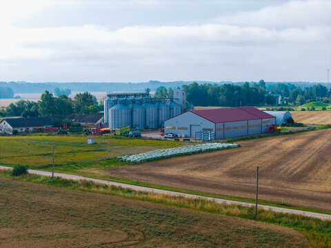 Aerial view of a farm featuring large grain silos, a modern warehouse with a red roof, and surrounding agricultural fields in Jaunberze, Dobele Municipality, Latvia.