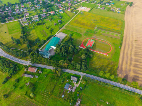 Aerial view of a community building with a green roof, a sports field with a red running track, and surrounding rural landscape in Jaunberze, Dobele Municipality, Latvia.