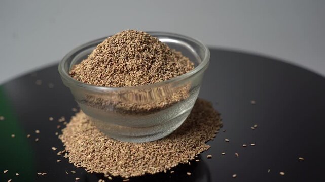 Close-up View of Carom Seeds in a Small Glass Bowl, Gently Rotate on a Black Reflective Surface