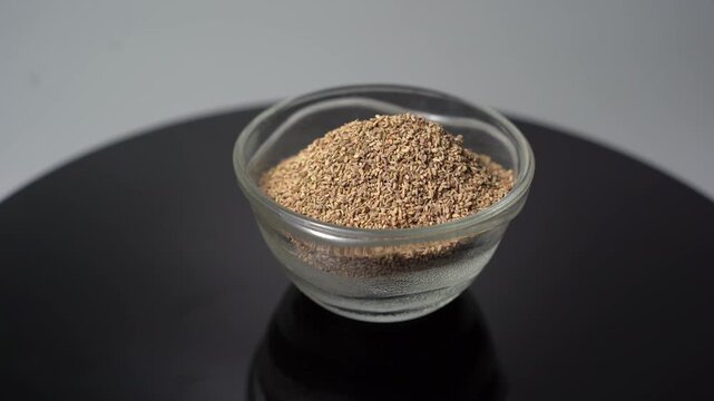 Ajwain Seeds (Carom Seeds) in a Glass Bowl on a Rotating Black Surface, Close-Up Shot with White Background