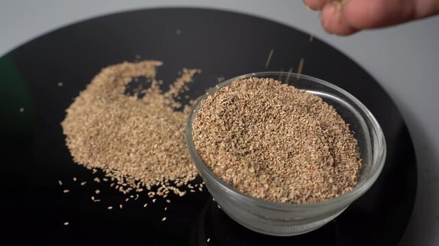 A close-up shot of a hand gently pour and sprinkle carom seeds on a black plate, with a glass bowl full of spices