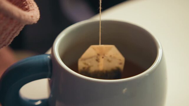 Tight close-up showing tea bag steeping in ceramic mug as amber liquid pours, drips back, and creates soft ripples across surface, with warm home kitchen light and quiet drink preparation