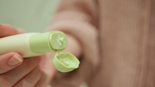 Close-up of hands preparing small pastel green-capped cylindrical stick applicator by twisting and removing cap to expose product tip; fingers grip tube and cap with gentle pressure while tube tilts