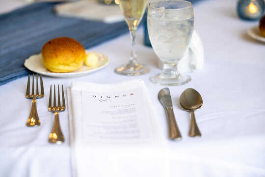 Elegant formal dinner place setting with menu, arranged flatware, water goblet, dinner roll, and refined tableware, illustrating fine dining, hospitality, entertaining, and special occasions.