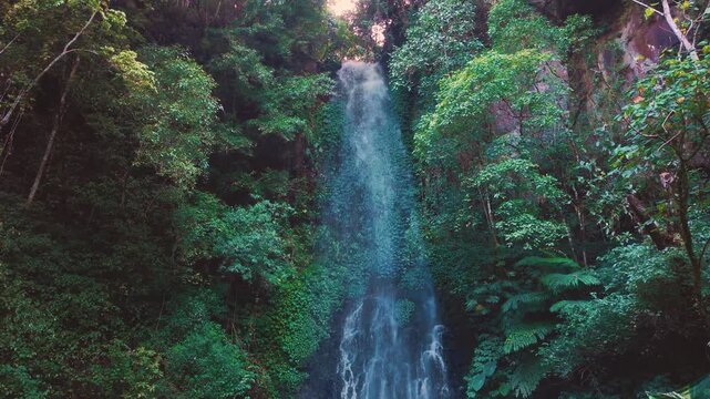 Aerial drone view of Coban Kaca waterfall flowing through dense tropical forest in Malang, East Java, Indonesia. The narrow waterfall cascades between lush green trees, creating a peaceful and seclude