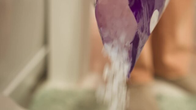White powder falling from purple packet into ceramic toilet bowl, with particles scattering through air and creating light splash and gentle ripples on water surface in indoor bathroom light