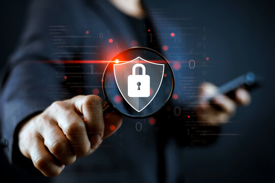 Business man holding magnifying glass with cyber security shield and padlock icon. Digital data protection, network safety, and information privacy concept on dark background.