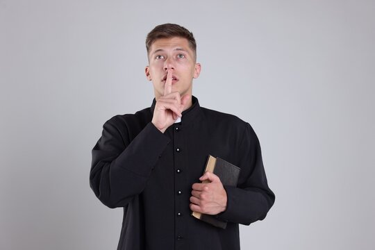 Priest in cassock with Holy Bible showing hush gesture on grey background