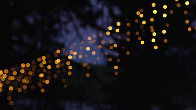 Warm string lights create golden bokeh among dark tree branches, forming a soft abstract night background with glowing circles of light and blurred foliage in a peaceful outdoor setting