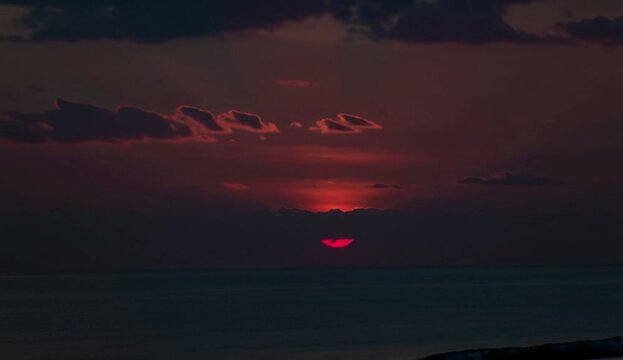 A dark, moody shot of a red sun partially obscured by clouds with hints of deep red and purple, a dark horizon waterline and a small silhouette in front. 4K video