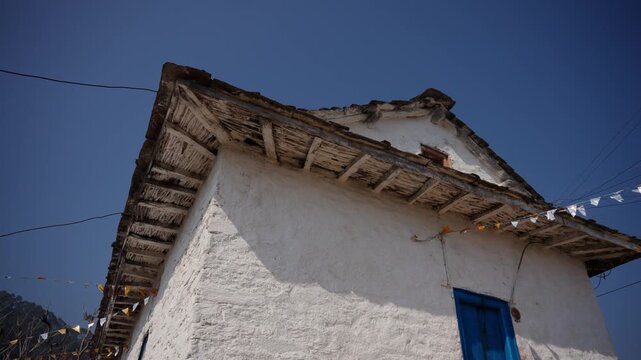 Low angle view of a whitewashed house roof corner with wooden eaves, a blue door below, and prayer flag bunting against a deep blue sky