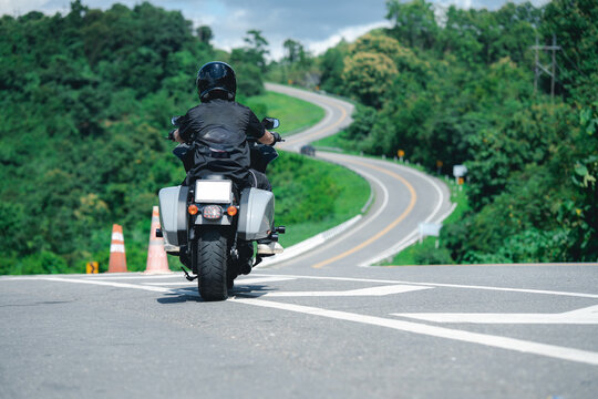 Motorcyclist wearing helmet rides along winding road surrounded by lush greenery and trees, evoking sense of adventure