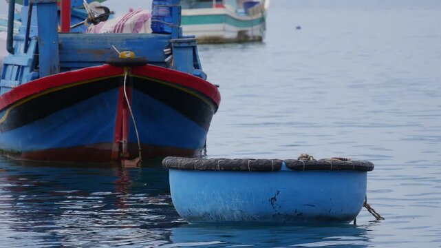 Small fishing boats float on calm water near the shore, with a blue boat in front and larger boats behind. Quiet harbor.