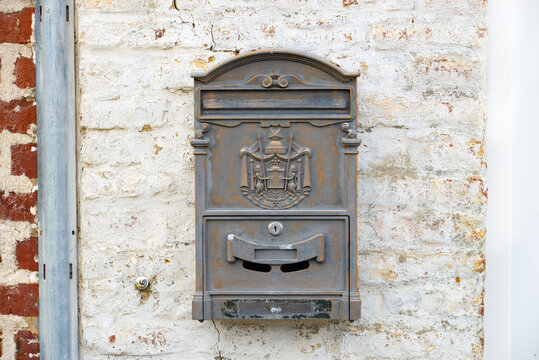 Old decorative metal mailbox mounted on a weathered white brick wall, featuring an ornate relief, letter slot, lock and aged rustic patina.