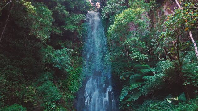 Aerial drone view of Coban Kaca waterfall flowing through dense tropical forest in Malang, East Java, Indonesia. The narrow waterfall cascades between lush green trees. 