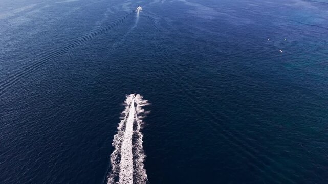A jet ski races across the open blue sea off Malta, leaving a long white wake, chased directly overhead by a forward-moving drone toward another vessel ahead.