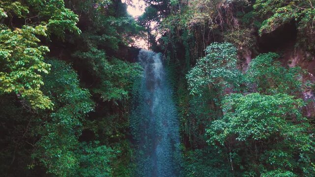 Aerial drone view of Coban Kaca waterfall flowing through dense tropical forest in Malang, East Java, Indonesia. The narrow waterfall cascades between lush green trees. 