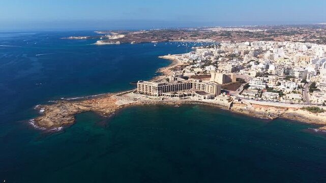 A wide aerial drone orbits and pulls back from the abandoned Jerma Palace hotel, revealing the coastal village of Marsaskala, Malta, and its rocky shoreline.