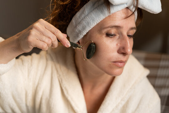 Spa wellness skincare ads. Woman in spa headband massaging face skin during daily skincare routine sitting on sofa using natural facial stone jade rolling device at home