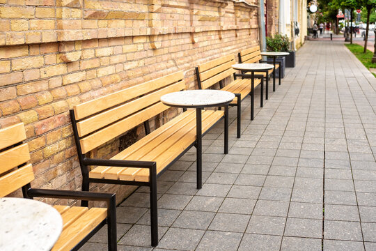 Empty outdoor cafe terrace with wooden benches and round tables along brick wall on sidewalk of city street receding into distance