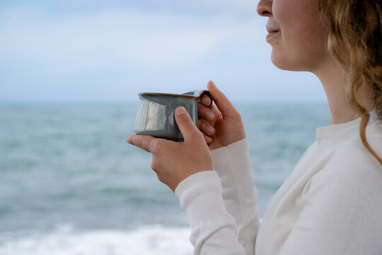 Cropped side view of a happy female holding a hot beverage near the ocean during cloudy daytime. The concept of relaxation and coastal vacation.