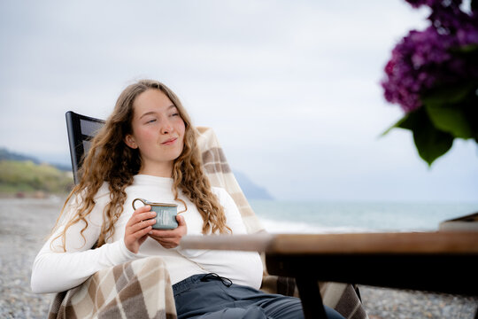 Woman enjoying a quiet moment with a warm drink on a pebble beach. Solitude, reflection and slow living by the seaside.