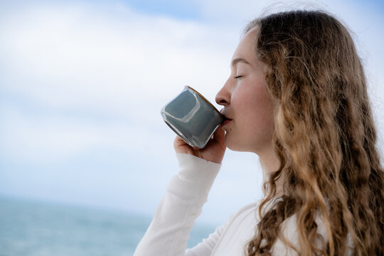 Curly woman savoring hot tea outdoors in nature with eyes closed near the ocean. Mindfulness, wellness and slow living concept.