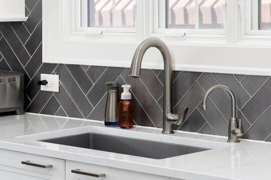 A kitchen faucet detail with a stainless steel faucet on a white marble countertop, white cabinets, and a dark grey textured herringbone tiled backsplash with white grout. 