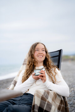 Smiling woman with a warm drink enjoying peaceful moments on the seashore, practicing slow living in loneliness, enjoying relaxation and self-care by the ocean.