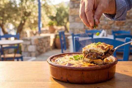 A steaming, rich slice of traditional Greek moussaka baked casserole with eggplant and bechamel sauce being served from a terracotta dish in an outdoor setting.