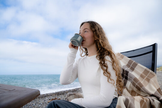 A young woman enjoying hot tea while relaxing by the ocean on a cloudy day. Girl enjoying her casual coastal lifestyle during cool daytime, with peaceful relaxation in nature and a slow travel concept
