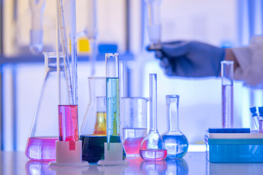 Chemistry laboratory scene with colorful chemical solutions in glass flasks and cylinders, scientist in gloves writing results, analytical experiment workspace.