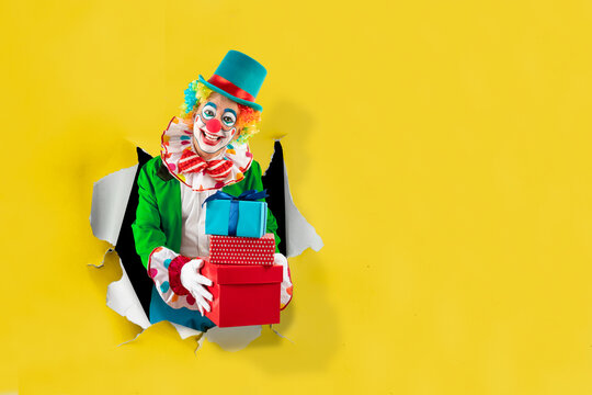 Funny clown with gifts looking through the ripped paper hole, man entertainer as Joker in a suit and wig. Trickster, jester, pantomime, mime, and a professional actor at events, birthday party.