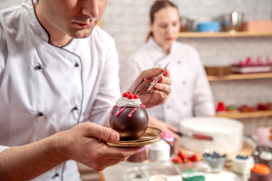 Pastry chef decorating an individual chocolate dessert with red berries and tweezers in a confectionery workshop